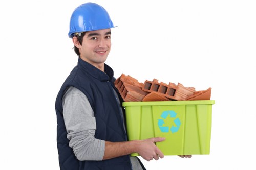 A young male waste collection worker wearing a blue safety helmet and a navy blue vest over a grey long-sleeved shirt, holding a green plastic recycling bin filled with red clay brick fragments and small blocks, set against a plain white background. The worker is smiling, with his right arm supporting the bottom of the bin and his left hand gripping the side, positioned slightly to the right of centre. The recycling bin features a blue recycling symbol on its front face. This image relates to rubbish removal and waste management services offered by Commercial Waste Removal Twickenham, serving local businesses and residents in Twickenham and nearby areas, including postcodes within the Twickenham region. The scene emphasizes an organized approach to waste disposal, focusing on construction or renovation debris, with the worker ready to transport the waste materials. The plain white backdrop ensures full focus on the individual, the bin, and the contents, illustrating a professional environmental service environment appropriate for a company's advertising for rubbish collection within the local London borough.