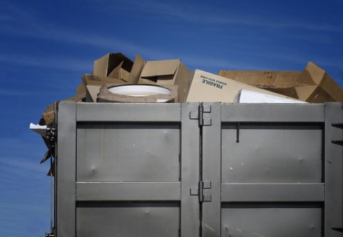 A large, open metal skip parked outdoors, filled with a variety of discarded materials including cardboard boxes, some flattened and others partially collapsed, alongside a few unidentifiable white objects. The cardboard exhibits a rough, corrugated texture with light brown and beige tones, and some of the boxes show visible printed labels. The skip is positioned on a paved surface, with the sky in the background displaying a clear, deep blue with minimal cloud cover. The surrounding environment suggests a typical waste collection site or commercial premises, aligning with rubbish removal services in Twickenham. The skip's metal surface appears slightly weathered, with subtle scratches and a matte finish, and the lid supports or handles are visible on the sides. The overall scene emphasizes the service provided by Commercial Waste Removal Twickenham for managing bulky waste and cardboard disposal in the local area.