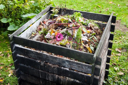 A black wooden compost bin filled with mixed organic waste, including decomposing plant material, wilted leaves, and some discarded flowers, situated outdoors on a grassy area near a garden bed. The compost bin is positioned on a patch of green grass with small scattered leaves around its base. In the background, there is greenery with additional plants and bushes, indicating a garden setting typical of residential areas in Twickenham. The scene appears to be taken during daylight hours, with natural light illuminating the waste materials and surrounding environment. This image is relevant to rubbish and waste management services provided by Commercial Waste Removal Twickenham, which specializes in removing green waste and organic debris from outdoor spaces.