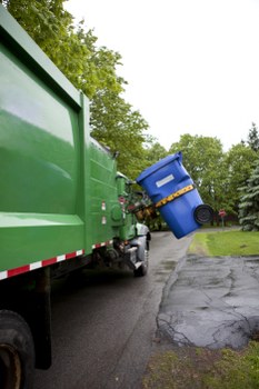 A large green waste collection truck parked on a residential street with trees and greenery in the background, positioned near the curb on the left side of the image. The truck’s rear container is visible, designed for rubbish collection, with a smooth, shiny surface and a reflective strip along its side. On the right side, a blue wheeled bin is being lifted or emptied into the truck’s hopper, with the bin tilted at an angle as it is positioned in the process of unloading. The street surface is asphalt with some cracks visible near the curb, and the sky appears overcast, suggesting a typical day for rubbish removal services in Twickenham. The scene is clean and organized, reflecting efficient waste collection activity by Commercial Waste Removal Twickenham, a company specializing in rubbish removal within the local area.