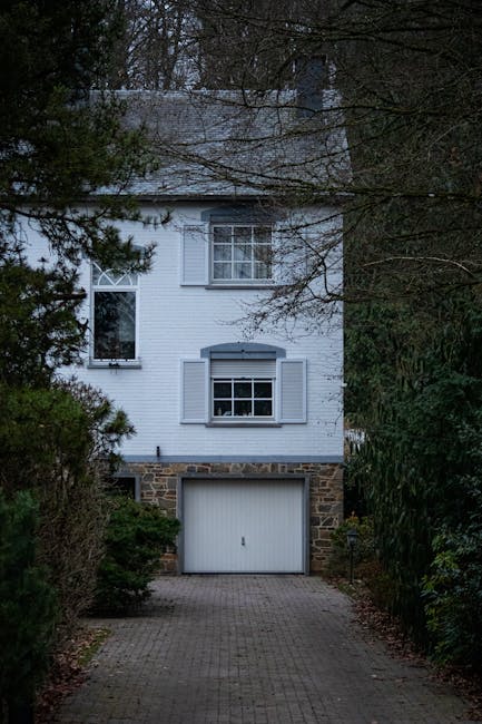 A three-storey detached house with a white exterior, metal roof, and shuttered windows, situated at the end of a paved driveway. The ground floor features a stone-clad garage door inset within a brick