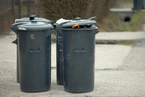 The image depicts two large, dark grey plastic wheelie bins positioned on a paved surface outdoors, likely in a residential or commercial area near Twickenham. The bin on the left has a closed lid, with a small, indistinct label affixed to the top, and a textured surface showing slight scuffs. The bin on the right has its lid partially open, revealing a glimpse of crumpled paper and an orange plastic object inside. The yard or driveway setting is visible in the background, with a blurred view of a concrete pathway extending into greenery and a small bridge structure in the distance. The scene is naturally lit, suggesting daytime, and the bins are typical of waste collection containers used for rubbish removal services. This setup aligns with the services provided by Commercial Waste Removal Twickenham, serving local businesses and residents in the area, including surrounding postcode districts in Twickenham and nearby areas.